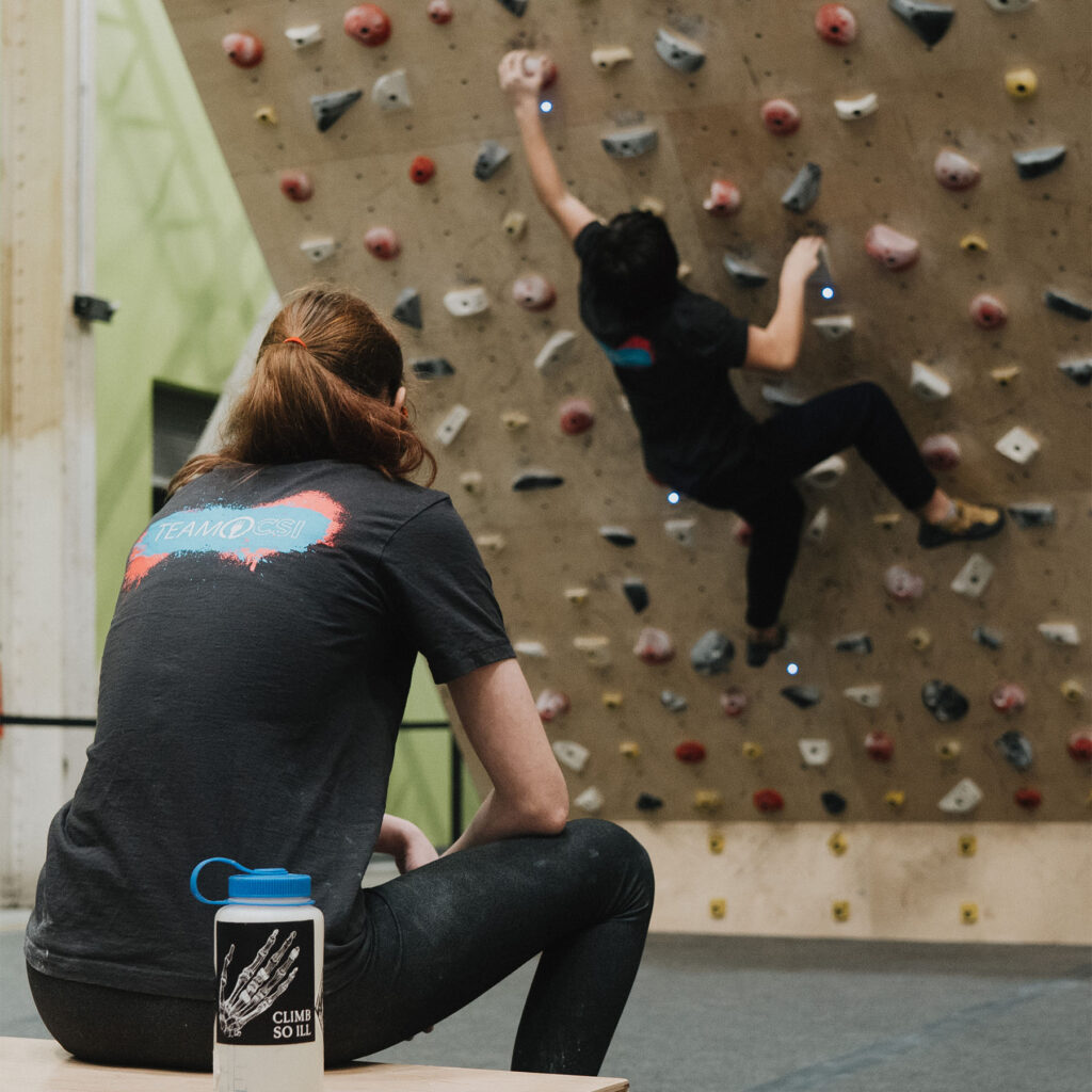 A climber trains on a Moon Board while another climber watches from a bench.